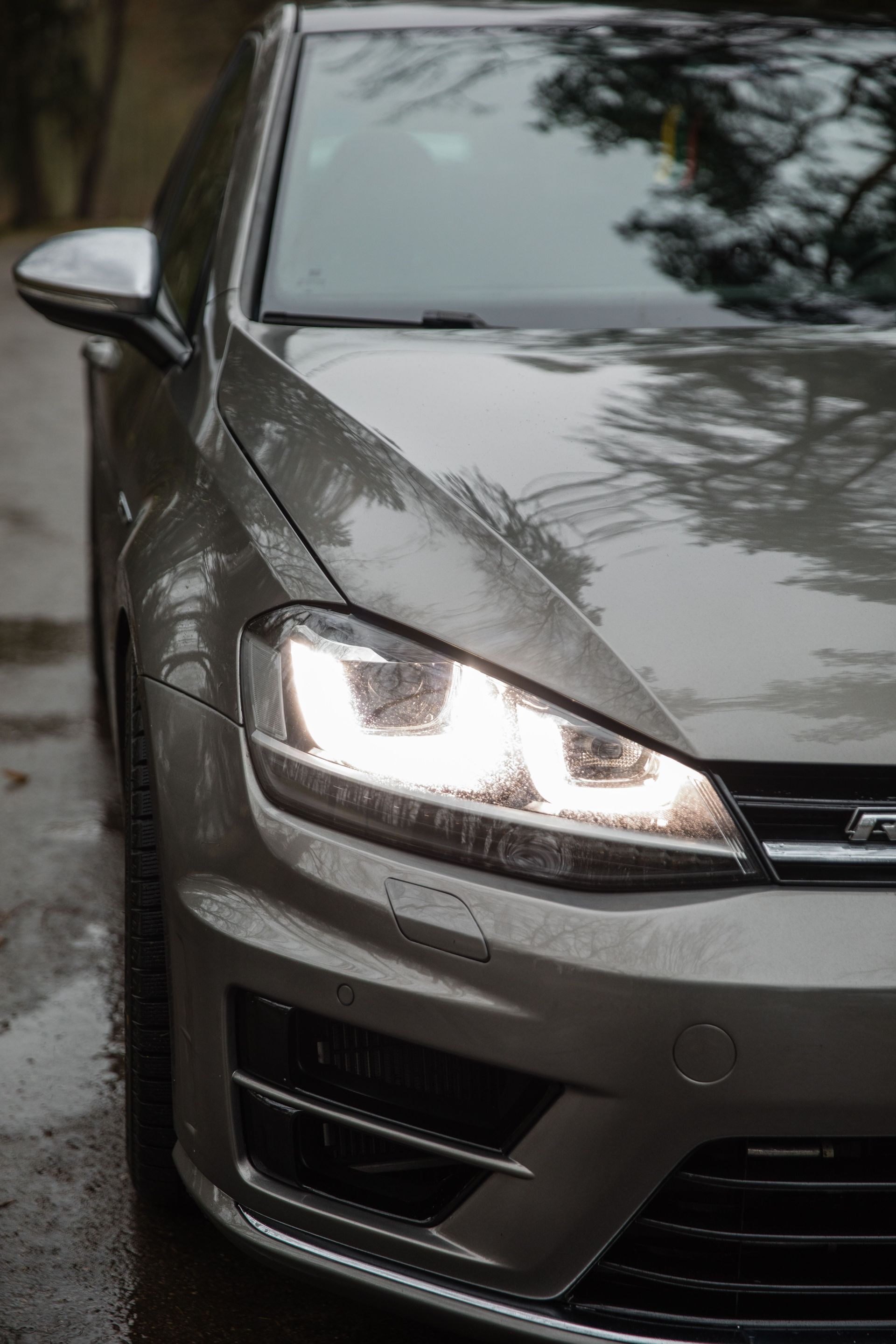 Close-up of a modern silver car with headlights on, reflecting nature.