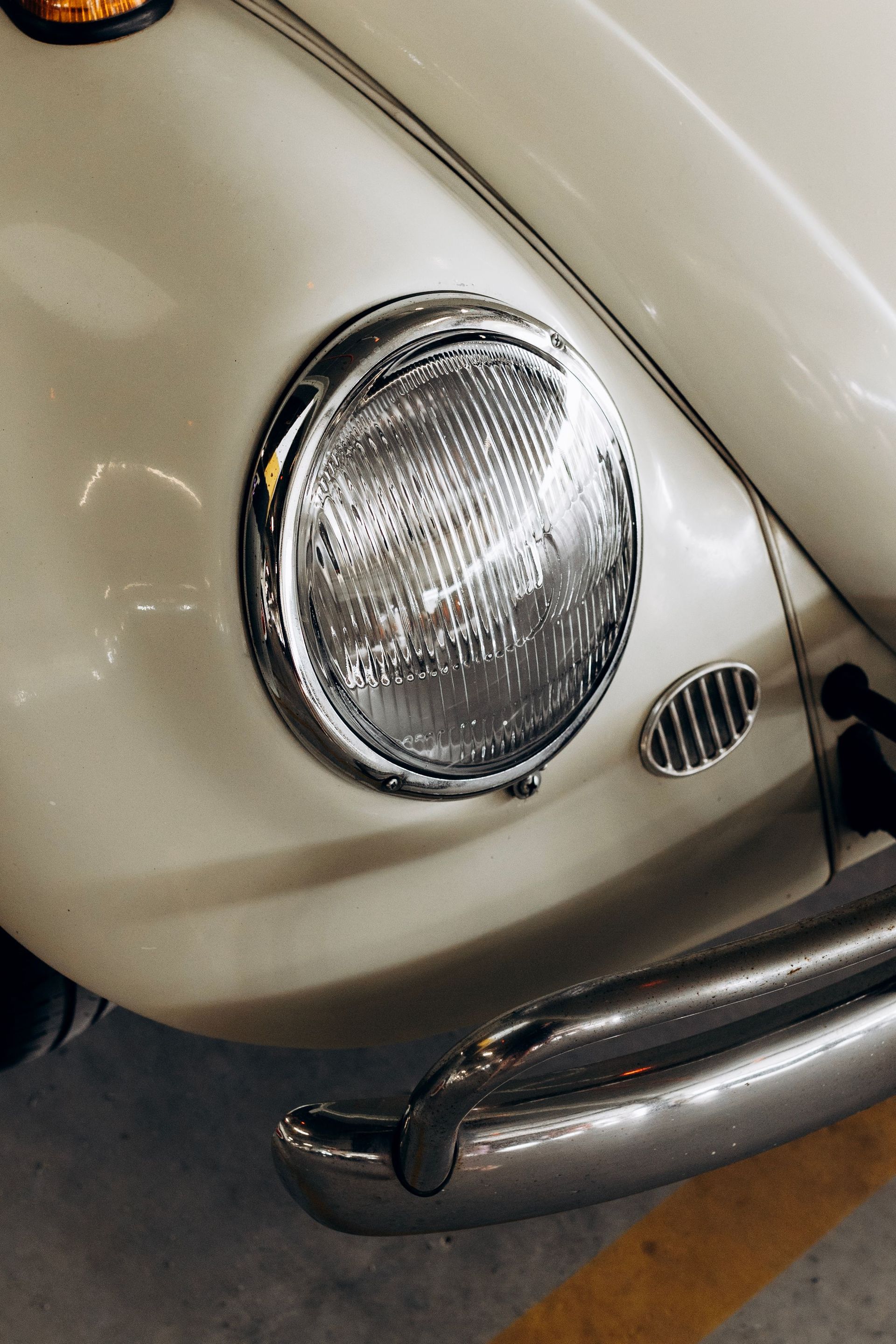 Detailed shot of a vintage white Volkswagen Beetle's headlight and bumper in a garage.